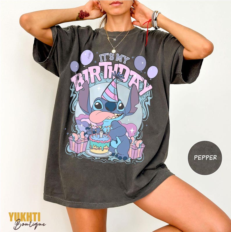 May include: Dark grey t-shirt with a cartoon character, birthday cake, and the text "IT'S MY BIRTHDAY". The design incorporates purple and pink balloons and gift boxes. The shirt is from Yukhti Boutique.
