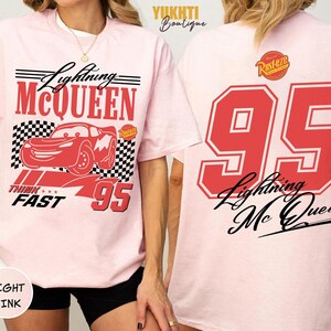 Puede incluir: Camiseta rosa claro con un dise&ntilde;o rojo de Lightning McQueen y el n&uacute;mero 95. El frente incluye "Lightning McQueen", "Think Fast" y un patr&oacute;n de bandera a cuadros. La parte trasera muestra un gran "95" rojo y "Lightning McQueen" en escritura negra.