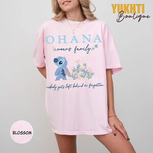 May include: A light pink t-shirt featuring the text "OHANA means family" with a cartoon character and floral accents. The phrase "nobody gets left behind or forgotten" is also printed on the shirt. The shirt has short sleeves.
