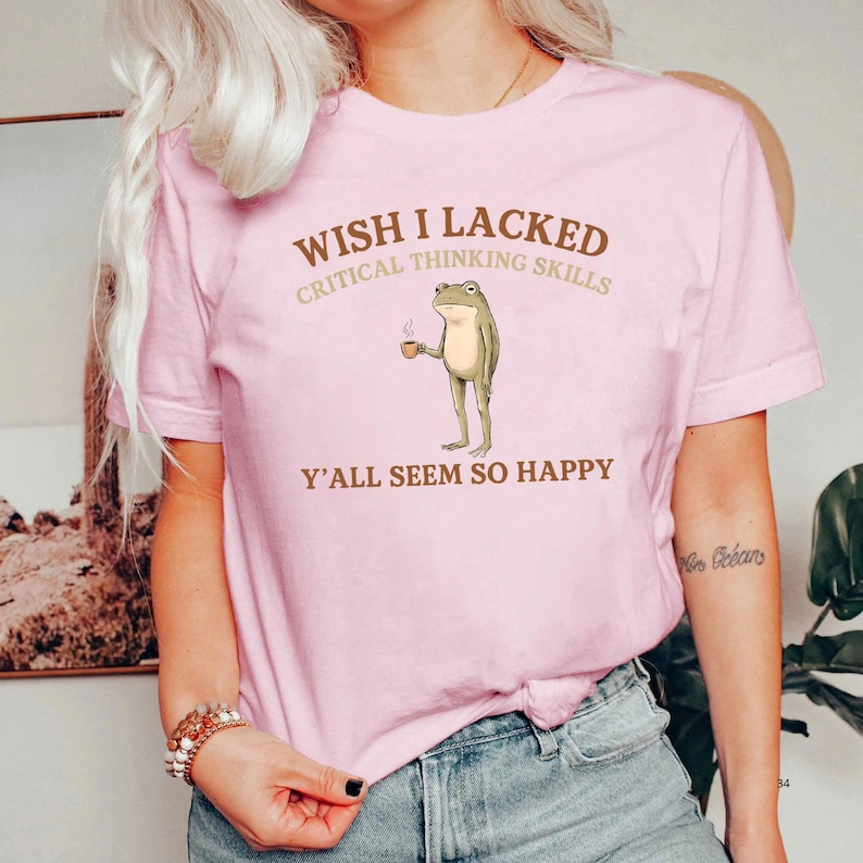 K&ouml;nnte beinhalten: Ein hellrosa T-Shirt mit kurzen &Auml;rmeln und einer Grafik eines stehenden Frosches, der eine kleine braune Tasse h&auml;lt. Der Text &uuml;ber dem Frosch lautet WISH I LACKED CRITICAL THINKING SKILLS und unter dem Frosch Y'ALL SEEM SO HAPPY.