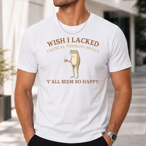 K&ouml;nnte beinhalten: Ein wei&szlig;es kurz&auml;rmeliges T-Shirt mit einer Grafik eines stehenden Frosches, der eine kleine braune Tasse h&auml;lt. Der Text &uuml;ber dem Frosch lautet WISH I LACKED CRITICAL THINKING SKILLS und darunter Y'ALL SEEM SO HAPPY in Braun- und Beiget&ouml;nen.