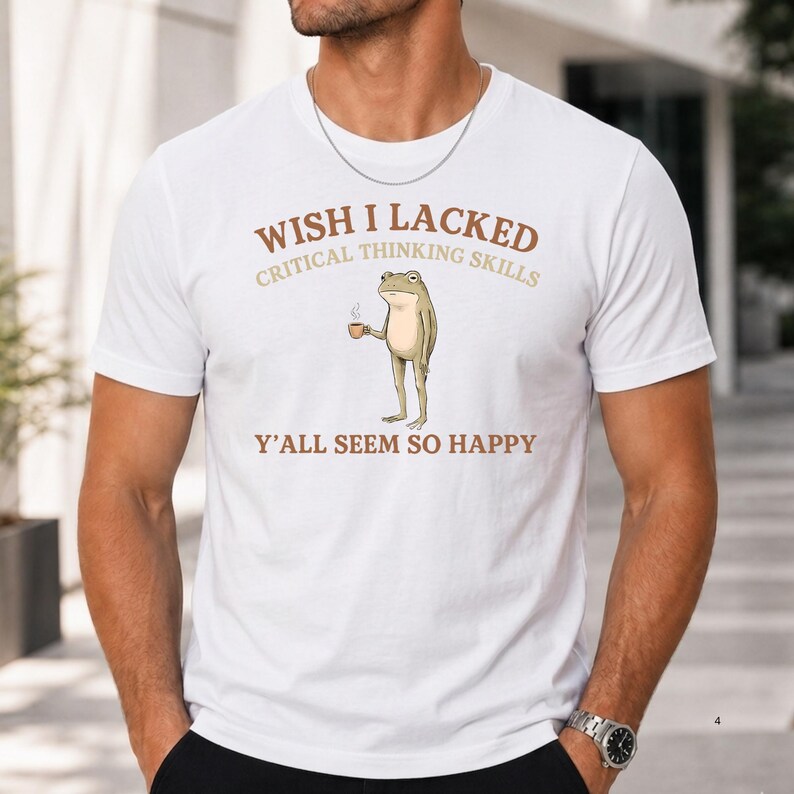 K&ouml;nnte beinhalten: Ein wei&szlig;es kurz&auml;rmeliges T-Shirt mit einer Grafik eines stehenden Frosches, der eine kleine braune Tasse h&auml;lt. Der Text &uuml;ber dem Frosch lautet WISH I LACKED CRITICAL THINKING SKILLS und darunter Y'ALL SEEM SO HAPPY in Braun- und Beiget&ouml;nen.