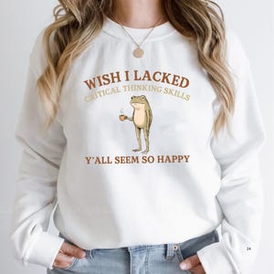 K&ouml;nnte beinhalten: Ein wei&szlig;es Langarm-Sweatshirt mit einer Grafik eines stehenden Frosches, der eine kleine braune Kaffeetasse h&auml;lt. Der Text WISH I LACKED CRITICAL THINKING SKILLS Y'ALL SEEM SO HAPPY ist in braunen und beigen Buchstaben auf der Brust gedruckt.