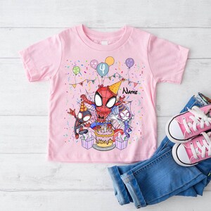 May include: Pink t-shirt featuring a Spider-Man birthday design. The graphic includes Spider-Man, a birthday cake, balloons, and confetti. The number "4" is displayed on a balloon. Blue jeans and pink sneakers are next to the shirt.