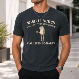 K&ouml;nnte beinhalten: Ein dunkel anthrazitfarbenes T-Shirt mit Rundhalsausschnitt und einer Grafik eines stehenden Frosches, der eine kleine Tasse h&auml;lt. Der Text lautet: WISH I LACKED CRITICAL THINKING SKILLS Y'ALL SEEM SO HAPPY. Das Design ist in Cremefarbe auf die Brust gedruckt.