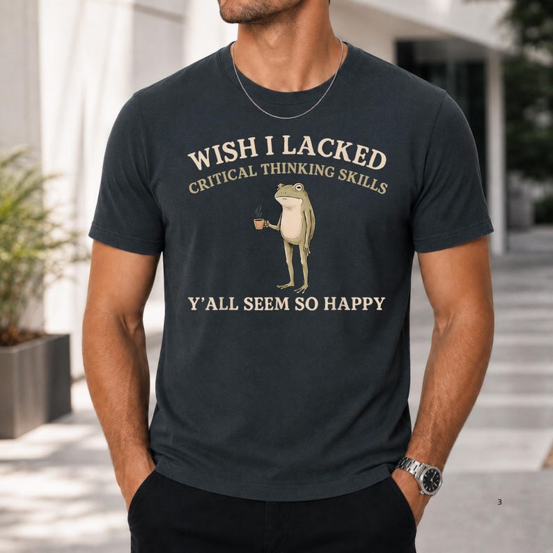 K&ouml;nnte beinhalten: Ein dunkel anthrazitfarbenes T-Shirt mit Rundhalsausschnitt und einer Grafik eines stehenden Frosches, der eine kleine Tasse h&auml;lt. Der Text lautet: WISH I LACKED CRITICAL THINKING SKILLS Y'ALL SEEM SO HAPPY. Das Design ist in Cremefarbe auf die Brust gedruckt.