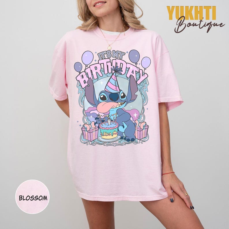May include: A pale pink t-shirt with a cartoon character and the text "IT'S MY BIRTHDAY". The design includes balloons, a birthday cake, and gift boxes. The shirt has short sleeves and a crew neck.