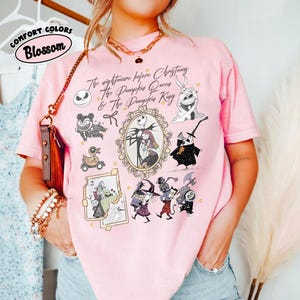 May include: Pink t-shirt with a graphic featuring characters and text from "The Nightmare Before Christmas." The design includes Jack and Sally in an ornate frame, along with other characters. The shirt also has the text "Comfort Colors Blossom."