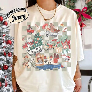 Puede incluir: Camiseta de color marfil con un gráfico colorido con personajes de dibujos animados con temática navideña. El diseño incluye un árbol de Navidad, un tren y varios personajes sobre un fondo a cuadros. Las palabras "COMFORT COLORS" e "Ivory" están impresas en la parte superior izquierda.