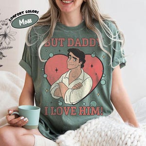 以下が含まれることがあります： モスグリーンのTシャツ。テキスト「BUT DADDY I LOVE HIM！」と赤いハートの中に漫画のキャラクターのグラフィック。シャツにはテキストの上に「COMFORT COLORS」という文字も入っています。