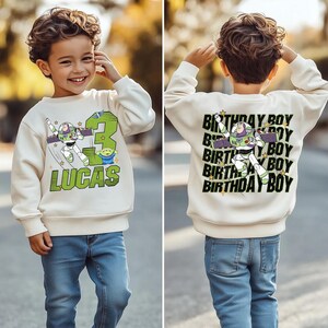 May include: A cream-colored sweatshirt featuring a Buzz Lightyear graphic, the number 3, and the name "LUCAS" in green. The back of the sweatshirt says "BIRTHDAY BOY" with a Buzz Lightyear graphic. The child is wearing blue jeans.