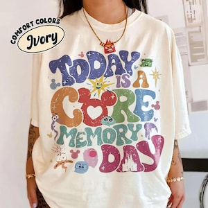 May include: Ivory Comfort Colors t-shirt with a colourful graphic reading "Today is a Core Memory Day." The design features cartoon characters and a Mickey Mouse-inspired heart. The shirt has a relaxed fit.