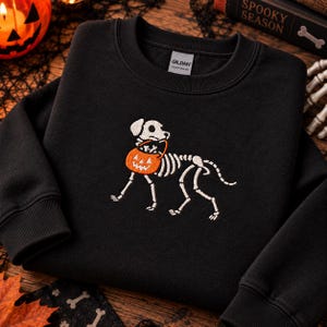 May include: A black crewneck sweatshirt featuring an embroidered design of a white dog skeleton carrying an orange jack-o'-lantern candy bucket in its mouth. The sweatshirt has a ribbed collar and cuffs, with a Gildan brand label at the neck.