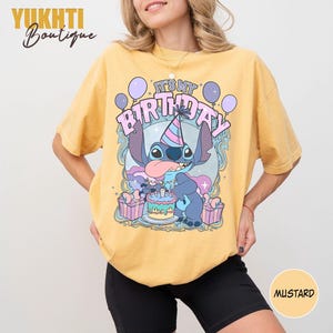 May include: Mustard yellow t-shirt with a cartoon character and the text "IT'S MY BIRTHDAY". The design includes a birthday cake, balloons, and gift boxes. The t-shirt has a loose fit.