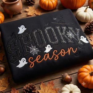 Op de afbeelding: Een zwart sweatshirt met de geborduurde tekst SPOOKY in grote zwarte glitterende blokletters en season in oranje cursief schrift. Het ontwerp bevat witte spoken, spinnenwebben, vleermuizen en een zwarte kat op een bezemsteel.