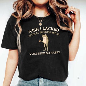 K&ouml;nnte beinhalten: Ein schwarzes Kurzarm-T-Shirt mit einer Grafik eines stehenden Frosches, der eine kleine braune Kaffeetasse h&auml;lt. Der Text &uuml;ber dem Frosch lautet WISH I LACKED CRITICAL THINKING SKILLS und der Text unter dem Frosch lautet Y'ALL SEEM SO HAPPY.