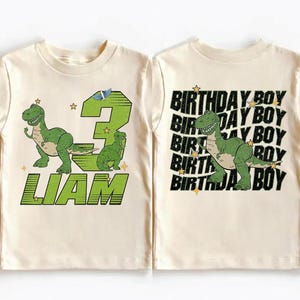 May include: A cream-colored toddler t-shirt featuring a green dinosaur graphic and the text "LIAM" in green. The number "3" is also displayed in green. The back of the shirt says "BIRTHDAY BOY" with a green dinosaur graphic.