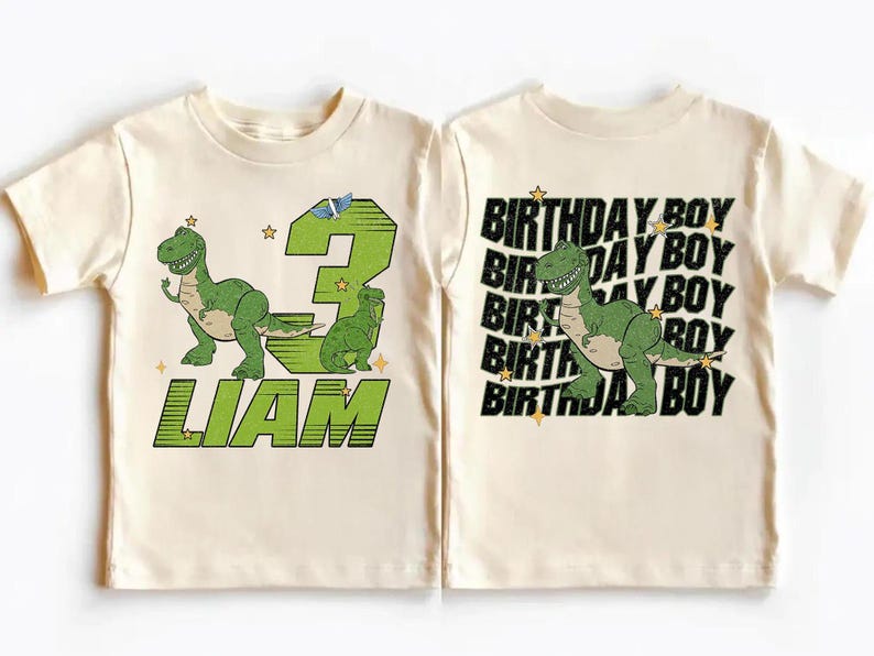 May include: A cream-colored toddler t-shirt featuring a green dinosaur graphic and the text "LIAM" in green. The number "3" is also displayed in green. The back of the shirt says "BIRTHDAY BOY" with a green dinosaur graphic.