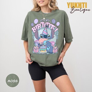 May include: Moss green t-shirt with a cartoon character and the text "IT'S MY BIRTHDAY". The design includes a birthday cake, balloons, and gift boxes. The shirt has a relaxed fit.