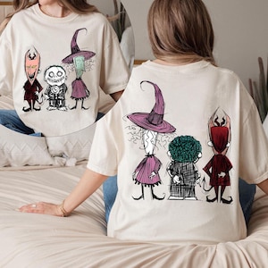 Pode incluir: Camiseta creme com um design de desenho animado caprichoso na frente e atrás. A frente mostra um diabo, um esqueleto e uma bruxa. A parte de trás mostra os mesmos personagens. A obra de arte é em preto, vermelho e roxo.