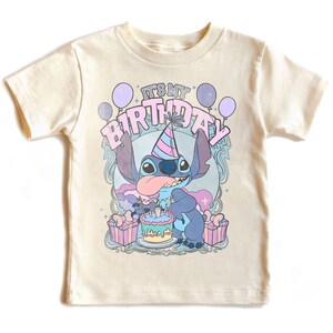 May include: A cream-coloured t-shirt with a cartoon character and the text "IT'S MY BIRTHDAY". The graphic includes balloons, a birthday cake, and gift boxes. The character is blue with a party hat and tongue sticking out.