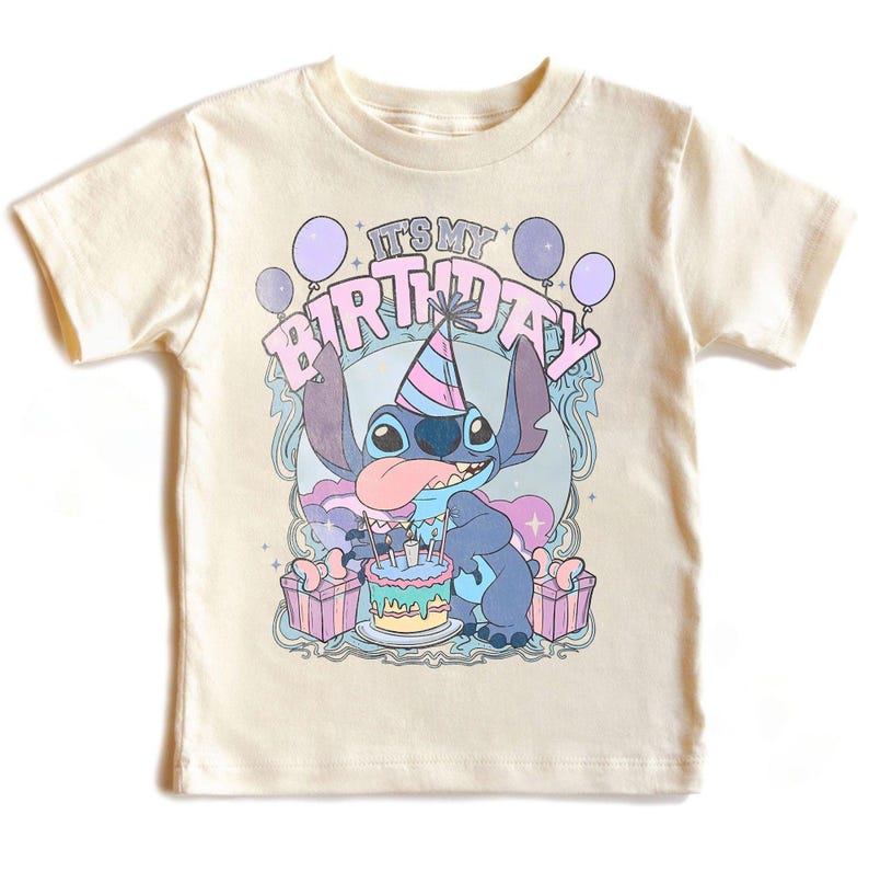 May include: A cream-coloured t-shirt with a cartoon character and the text "IT'S MY BIRTHDAY". The graphic includes balloons, a birthday cake, and gift boxes. The character is blue with a party hat and tongue sticking out.