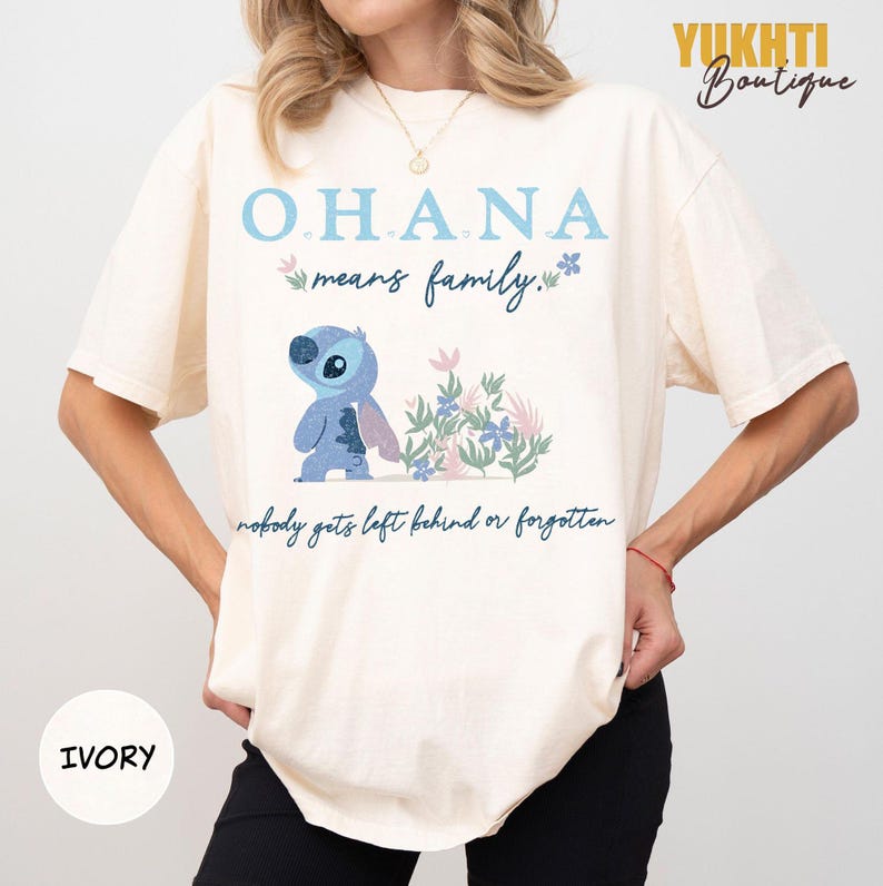 May include: Ivory-colored t-shirt featuring the text "OHANA means family" in light blue and a cartoon character. The design includes floral accents and the phrase "nobody gets left behind or forgotten."
