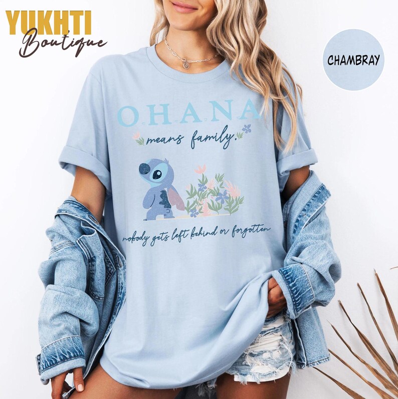 May include: Light blue t-shirt with the word "OHANA" and the phrase "means family" in light blue. A cartoon character is featured below the text, with the phrase "nobody gets left behind or forgotten" at the bottom. The shirt is chambray.