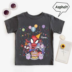 May include: Dark gray t-shirt featuring a colorful graphic of Spider-Man and two other Spider-Man characters celebrating a birthday. The design includes a cake, balloons, confetti, and the number 4. The word "Name" is also visible.