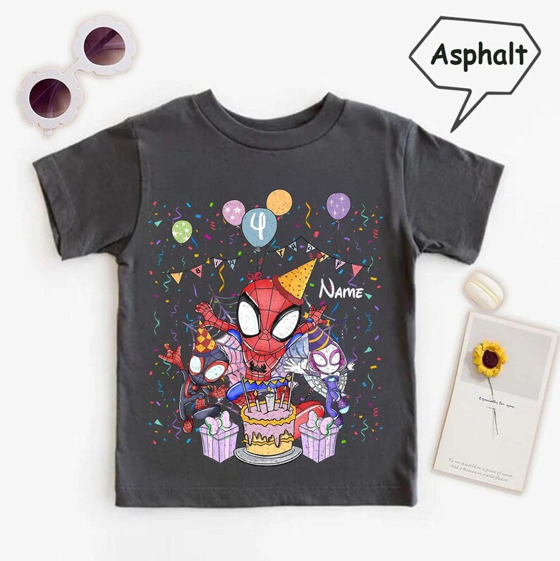 May include: Dark gray t-shirt featuring a colorful graphic of Spider-Man and two other Spider-Man characters celebrating a birthday. The design includes a cake, balloons, confetti, and the number 4. The word "Name" is also visible.