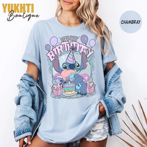May include: Pale blue t-shirt with a cartoon character celebrating a birthday. The graphic includes the text "IT'S MY BIRTHDAY" with balloons, a birthday cake, and wrapped gifts. The shirt is a chambray colour.