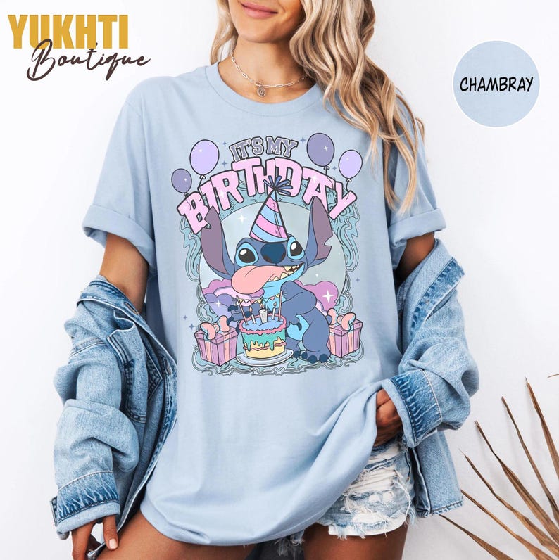 May include: Pale blue t-shirt with a cartoon character celebrating a birthday. The graphic includes the text "IT'S MY BIRTHDAY" with balloons, a birthday cake, and wrapped gifts. The shirt is a chambray colour.