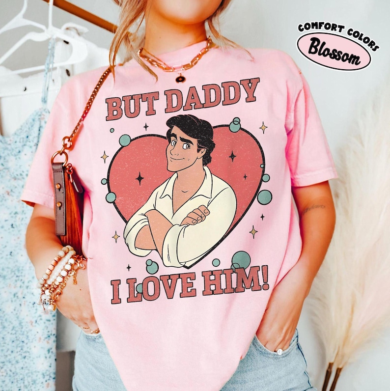 以下が含まれることがあります： 「BUT DADDY I LOVE HIM！」の文字と、ハートの中に漫画の男性が描かれたピンク色のTシャツ。「COMFORT COLORS Blossom」の文字も円で囲まれています。