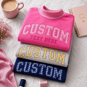 May include: Three stacked sweatshirts with mock neck collars. The top is hot pink with pink gingham lettering reading CUSTOM TEXT HERE. The middle is heather grey with yellow gingham lettering. The bottom is navy blue with blue gingham lettering.