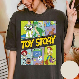 May include: Dark gray t-shirt featuring a colorful graphic of characters from the animated film "Toy Story." The design includes Buzz Lightyear, Rex, Woody, and the Little Green Men, with the words "Toy Story" in yellow.