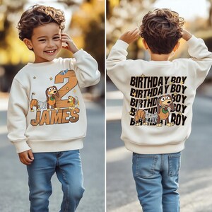 May include: Cream-colored sweatshirt with a cartoon dog and the number 2, with the name "JAMES" in a brown font. The back of the sweatshirt says "BIRTHDAY BOY" multiple times with a cartoon dog. The child is wearing blue jeans.