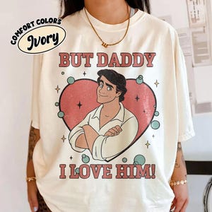 以下が含まれることがあります： 「BUT DADDY I LOVE HIM！」の文字と、ピンクのハートの中に男性の漫画イラストが描かれた、アイボリーのComfort Colors Tシャツ。クルーネックと半袖です。