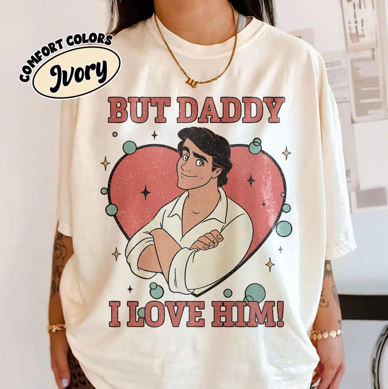 以下が含まれることがあります： 「BUT DADDY I LOVE HIM！」の文字と、ピンクのハートの中に男性の漫画イラストが描かれた、アイボリーのComfort Colors Tシャツ。クルーネックと半袖です。