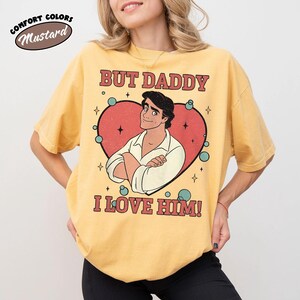 以下が含まれることがあります： マスタード色のTシャツ。テキスト「BUT DADDY I LOVE HIM！」と赤いハートの中の男性の漫画イラストが描かれています。シャツには「COMFORT COLORS」と「Mustard」という言葉も円で表示されています。