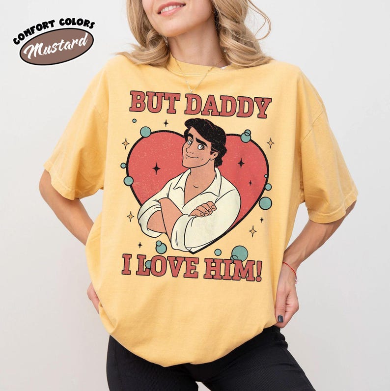 以下が含まれることがあります： マスタード色のTシャツ。テキスト「BUT DADDY I LOVE HIM！」と赤いハートの中の男性の漫画イラストが描かれています。シャツには「COMFORT COLORS」と「Mustard」という言葉も円で表示されています。