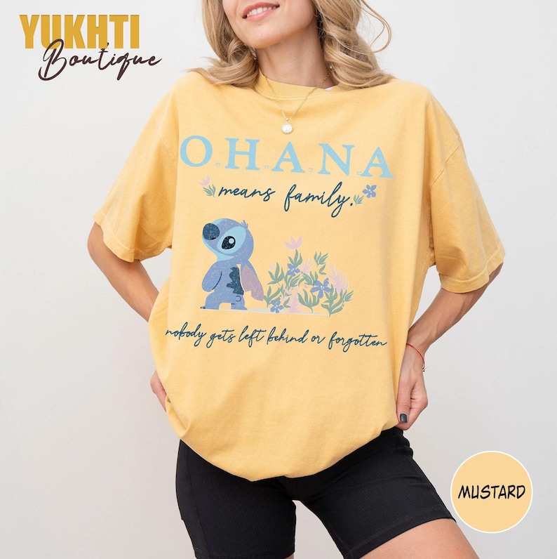 May include: Mustard yellow t-shirt featuring the text "OHANA means family" with a cartoon character and floral accents. The shirt also includes the phrase "nobody gets left behind or forgotten."