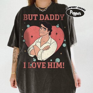 以下が含まれることがあります： 「BUT DADDY I LOVE HIM！」の文字がプリントされた濃いグレーのTシャツ。ピンクのハートの中に腕を組んだ男性の漫画イラストが描かれています。シャツには白い楕円の中に「COMFORT COLORS Pepper」の文字も入っています。
