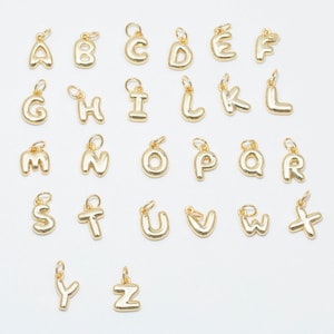 May include: A selection of gold-coloured alphabet charms, each with a small loop for attaching to jewellery. The letters are in a rounded, bubble font, and are arranged in rows on a white surface. The charms are all the same size and style.