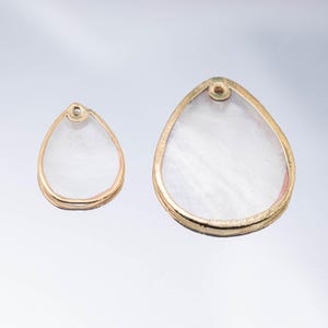 May include: Two teardrop-shaped pendants. They have a gold-coloured frame and a pearlescent, white interior. The pendants are of different sizes, with a small hole at the top for stringing. The gold frame has a textured appearance.