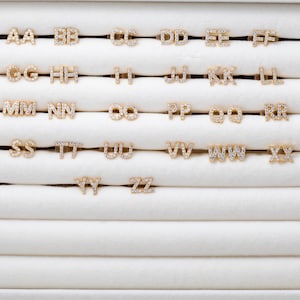 Può includere: Una collezione di orecchini a lobo con iniziali color oro, ciascuno incastonato con piccoli cristalli scintillanti. Le lettere AA-ZZ sono visualizzate in una griglia, mostrando la varietà di opzioni di gioielli personalizzati disponibili. Gli orecchini sono presentati su una superficie bianca strutturata.