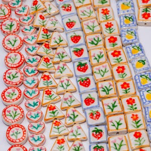 May include: A selection of ceramic beads in diverse shapes: circles, hearts, triangles, and squares. The beads are adorned with hand-painted floral, strawberry, and lemon motifs. The colour scheme features red, pink, green, yellow, and blue, creating a vibrant display.