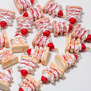 May include: A collection of miniature cake charms with a strawberry accent. The charms feature layers of tan-colored cake, white frosting, and red strawberry filling. Each charm has a gold-colored loop for attaching to jewelry or other crafts.