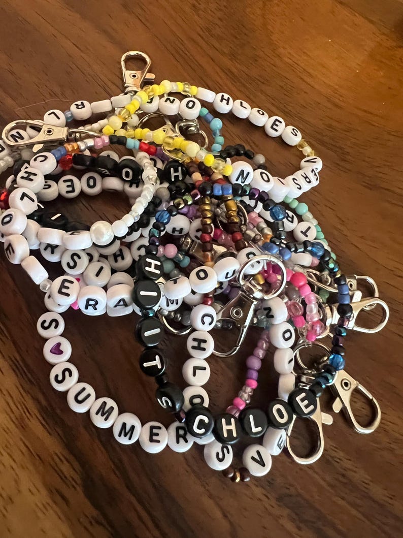 May include: A collection of beaded keychains with various names spelled out in black and white letter beads. The keychains feature silver-colored clasps and a variety of colorful beads. Some keychains spell out names like "SUMMER" and "CHLOE".