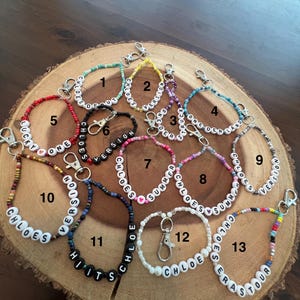 May include: A collection of beaded keychains with various colors and text. The keychains feature white letter beads spelling out names and phrases like "Chloe's Version", "Summer Chloe's", and "Hits". Each keychain has a silver clasp.