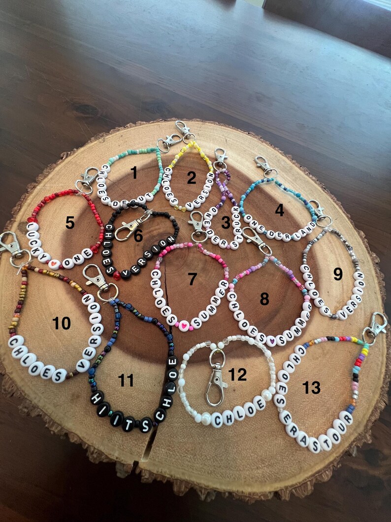 May include: A collection of beaded keychains with various colors and text. The keychains feature white letter beads spelling out names and phrases like "Chloe's Version", "Summer Chloe's", and "Hits". Each keychain has a silver clasp.